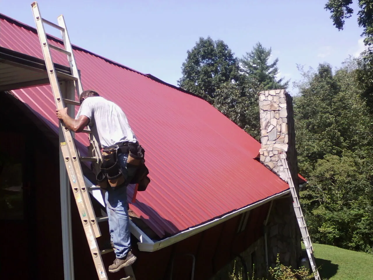 Roofer on a ladder beside a red metal roof and stone chimney — Soffit & Fascia Repair by Roofing Brothers in Olmsted Falls